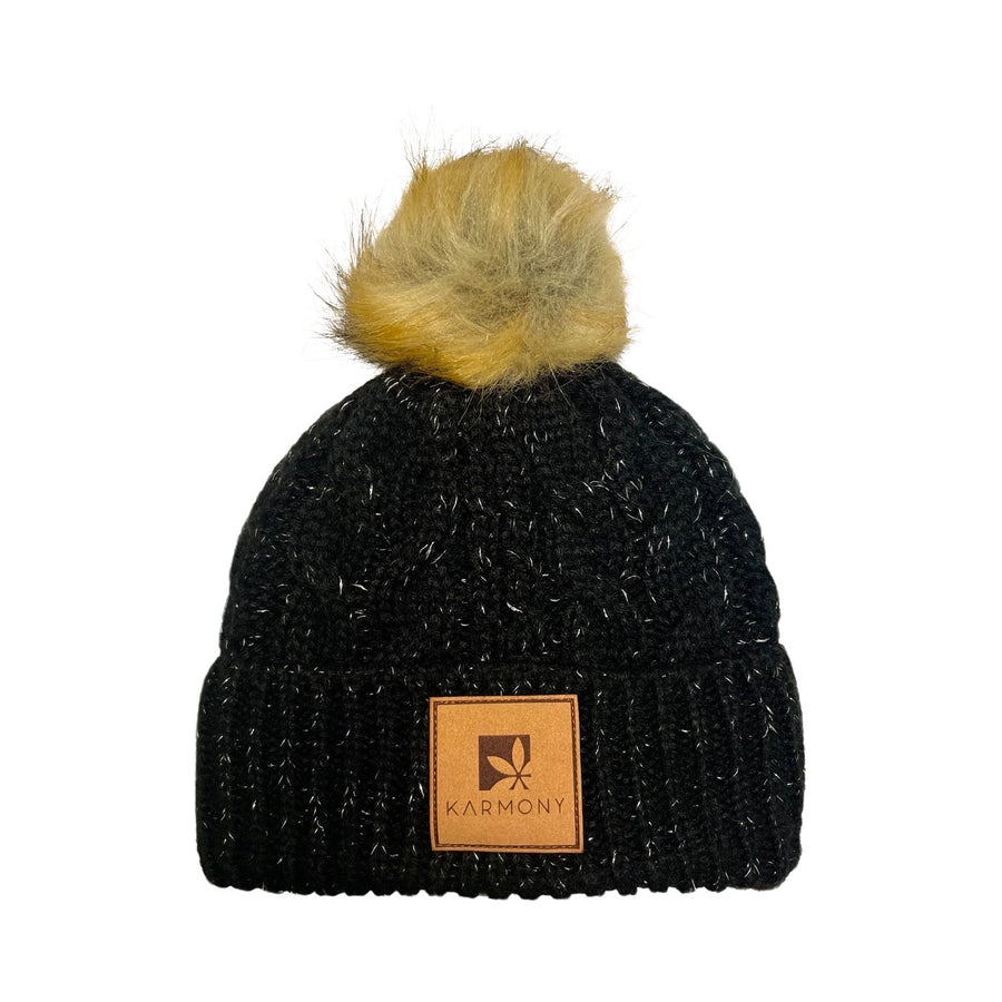 Black beanie with silver metallic flecks and brown faux fur pompom. The brim has a leather patch of the Karmony logo.