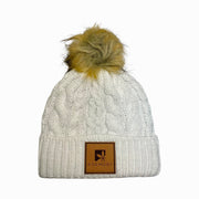 Cream beanie with silver metallic flecks and brown faux fur pompom. The brim has a leather patch of the Karmony logo.