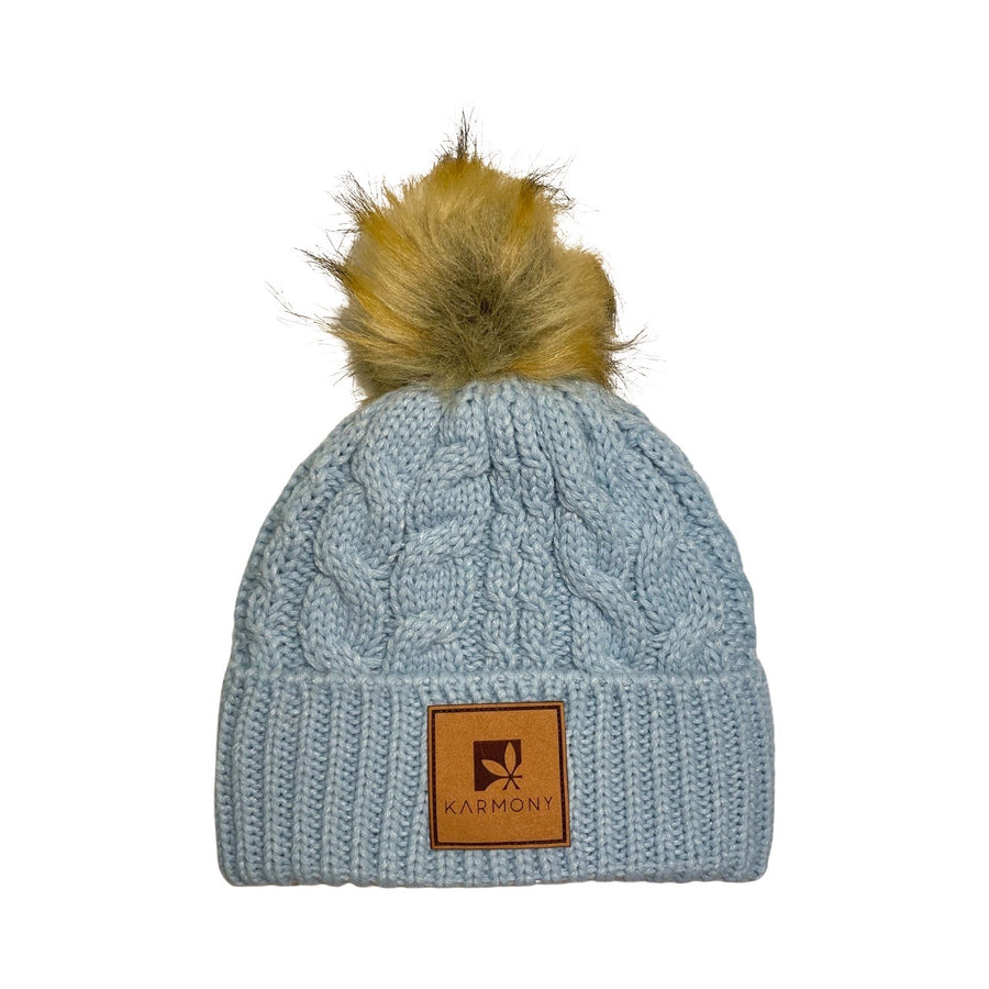 Sky blue beanie with silver metallic flecks and brown faux fur pompom. The brim has a leather patch of the Karmony logo.
