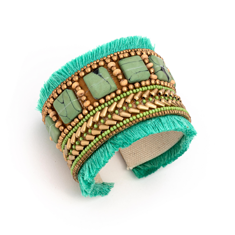 A thick cuff bracelet with tan, green, and turquoise beaded designs, and turquoise fringe.