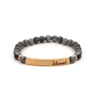 A bracelet of marbled grey and black stone beads, with a gold rectangular bar that reads "blessed".